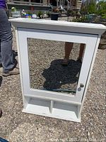 White medicine cabinet with mirror on door, photographed closed on gravel surface with person's legs visible.