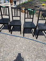Four black wooden chairs arranged outdoors on gravel, showing fronts and backs with visible wear and dirt.