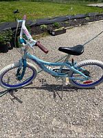 Left side profile of light blue Dyno single speed bike with purple rims and white tires, black seat, and brown worn grips on handlebars. Standing on gravel surface.