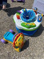 Overall lot showing LeapFrog ExerSaucer and Fisher-Price Lion walker on outdoor gravel surface with child feet visible nearby.