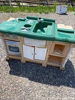Front view of the children's outdoor play kitchen showing green molded sink, faucet, stove knobs, white oven door, double cabinet doors, and open shelving underneath.