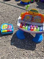 Two children's toy pianos outdoors on gravel, Fisher Price piano on the right with blue legs and multicolored keys, Little Tykes smaller piano on the left.