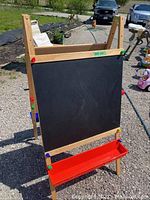 Wooden easel front side with chalkboard and red tray for chalk and accessories, shown outside on gravel.