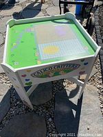 Front angle view of the white wooden Calico Critters activity table with illustrated tabletop and drawer, outdoors on stone pathway.