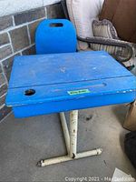 Front view of the blue school desk showing the top surface with worn paint, attached blue chair, hole for inkwell, and white metal base.