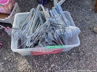 Plastic bin filled with many assorted metal wire coat hangers, silver color, placed outdoors on gravel surface.