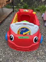 Red and yellow plastic Fisher Price activity center with blue wheels in outdoor setting on ground with gravel and grass.
