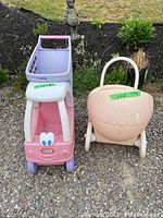 Photo showing front and top view of Pink and lavender Little Tikes shopping cart and pink doll stroller side by side on gravel surface with garden background.