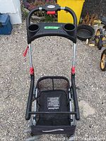 Front view of Baby Trend Snap-N-Go stroller frame showing handle with two cup holders and lower storage basket. Frame shows rust spots on metal parts.