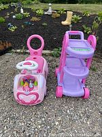 Pink Disney ride on toy with princess decorations and heart-shaped steering wheel alongside purple and pink three-tier plastic tea cart on gravel ground.