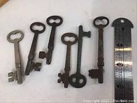Seven old skeleton keys arranged in one line with a ruler for size reference, showing varying lengths, metal material and patina.