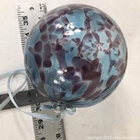 Image showing hand blown glass ball with purple and blue spotted pattern and a translucent hanging loop, placed next to rulers for scale.