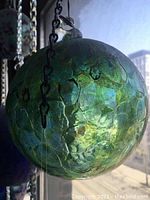 Close-up of hand blown glass ball showing green-blue hues and textured surface hanging by a black metal chain near window