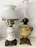Full frontal photo of two vintage hand painted lamps side by side. One is white hobnail milk glass with floral hand painting and the other is a round base lamp with pink roses and green foliage with a clear glass chimney. Both rest on ornate metal bases.