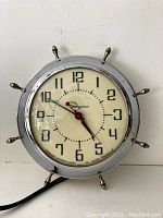 Front view of the vintage Ingraham wall clock showing its chrome ship wheel style frame and cream clock face with black numerals and red hands.