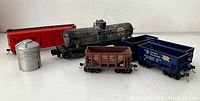 A photo of six toy train cars including a black tanker, several hopper cars, and a red Burlington boxcar alongside a grey cylindrical metal piece.