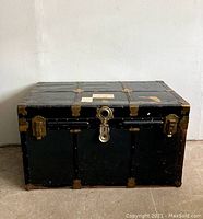 Full front view of vintage metal trunk showing black surface, brass corner protection, central lock, and two side latches.