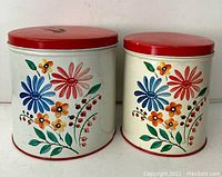 Two vintage metal canisters with white bodies decorated with colorful hand-painted style flowers and red lids shown side by side.