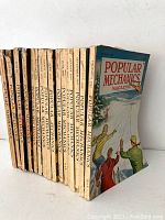 Photo of 16 vintage Popular Mechanics magazines stacked vertically showing covers and spines.