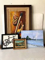 Four art pieces displayed against a wall: a large framed textile piece with fishing items, a small lighthouse landscape painting, a black framed bicycle illustration, and a small framed bridge print.