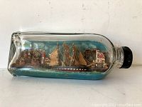 Clear glass bottle with a miniature ship and coastal village scene inside, viewed from side on a white background.