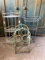 Three metal racks placed on floor against wooden backdrop - white corner shelf, gray basket rack, and green bottle rack.