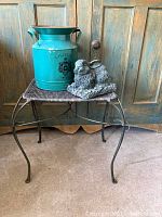 Photo of iron table with wicker top, teal tin bucket, and resin rabbit on top in front of wooden cabinet.