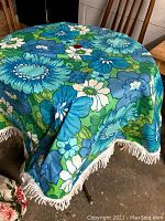 Blue floral patio table cloth with white fringe draped on a round table, showing vibrant blue and green mid century modern style floral pattern.