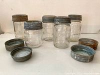 Five vintage glass canning jars with metal lids removed and placed in front, showing condition and details of lids and jars.