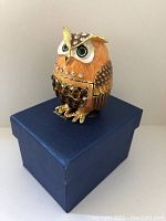 Owl-shaped trinket box sitting closed on a blue box, showing front and side profile with rhinestone embellishments and enamel finish.