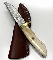 Hunting knife laid atop a brown leather sheath showing the bone handle, patterned steel blade, and brass guard