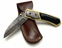Side view of open folding Damascus steel blade pocket knife with brass and black handle, resting on brown leather sheath with stitched edge and snap closure.