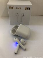 Photo showing white i9S-TWS Bluetooth earbuds outside of the charging case, glowing indicator light visible on earbuds and open case, box in background.