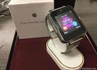 Smartwatch showing clock interface with date and time, displayed on white charging dock, box in background