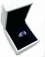 Ring with rectangular deep blue stone in center and pear-shaped clear stones on sides, placed in white jewelry box with black velvet interior.