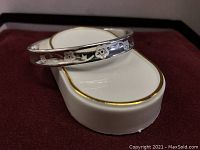 Image showing the sterling silver bangle resting on a white and gold-trimmed cushion, highlighting the plum flower engraving on the outer surface.