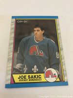 Front of 1989 O-Pee-Chee Joe Sakic rookie card featuring player in blue Quebec Nordiques jersey with team logo.