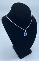 The mounted blue teardrop-shaped stone pendant on a silver twist link chain displayed on a black bust stand.