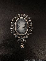 Front close-up of gray cameo brooch showing woman's profile and black stone setting on dark background.