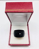 Black titanium ring displayed in a red and white ring box showing the front and inside view, highlighting the ring's band width and polished finish.