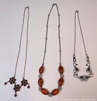 Three sterling silver necklaces laid flat: one with amber stones, one with dark pink clustered glass stones, one with blue and white crystals.