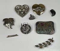 Top view showing all eight items: heart-shaped silver bird brooch, gold-toned heart bird brooch, leaf pin, circular floral brooch, small heart brooch, bear abalone/mother of pearl inlay brooch, curved branch with mother of pearl leaves brooch, and small bird earring.
