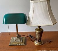 Two lamps displayed side by side on wood surface against grey wall: green banker desk lamp left, tall brass table lamp with fabric shade right