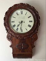 Front view of carved wooden wall clock with white enamel dial, Roman numerals, maker's mark and carved wreath motif