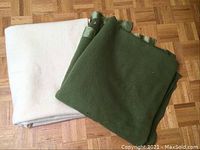 Photo showing the two folded wool blankets on a parquet wood floor. One is green with satin trim, the other beige without a visible tag.