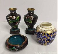 Photo showing two black mini urns with floral and brass details, one floral ashtray and a white vase with gold and blue design.