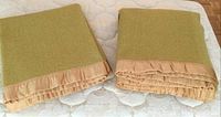 Two folded mustard-green wool blankets with peach satin trim placed side-by-side on a mattress with quilted cover, showing condition and color.