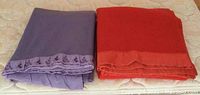 Two folded wool blankets side by side on mattress, purple with floral trim on left, red plain on right.