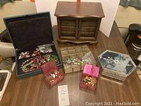View of all jewelry boxes with various costume jewelry pieces displayed on the table: wooden jewelry box, blue jewelry case, red small boxes, plastic compartmentalized tray, and hexagonal silver-tone jewelry box with green interior.