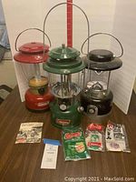 Three Coleman lanterns on a wooden table with accessories and manuals in front. Red electric lantern, green fuel lantern, and black Max lantern.